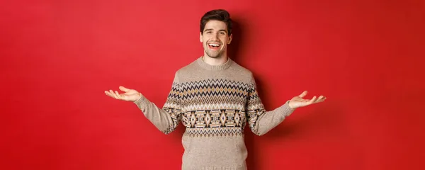 Portret van een gelukkig knappe man die nieuwjaarsfeestdagen viert, kersttrui draagt, handen zijwaarts spreidt en glimlacht, iets vasthoudt op kopieerruimte, over rode achtergrond staat — Stockfoto