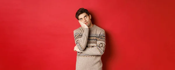 クリスマスのセーターに身を包んだ若い男のイメージ,ロール目と手にもたれ,喜びと刺激を表現します,赤い背景の上に立って — ストック写真