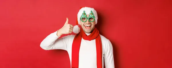 Noel, kış tatili ve kutlama kavramı. Noel Baba şapkası ve parti gözlüğü takmış neşeli bir adam. Başparmak kaldırıyor ve gülümsüyor. Kırmızı arka planda dikiliyor. — Stok fotoğraf