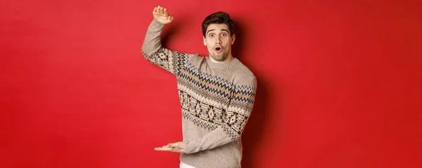 Imagen de un hombre atractivo en jersey de Navidad, dando forma a un gran regalo de año nuevo, mostrando algo grande y sorprendente, de pie sobre el fondo rojo —  Fotos de Stock