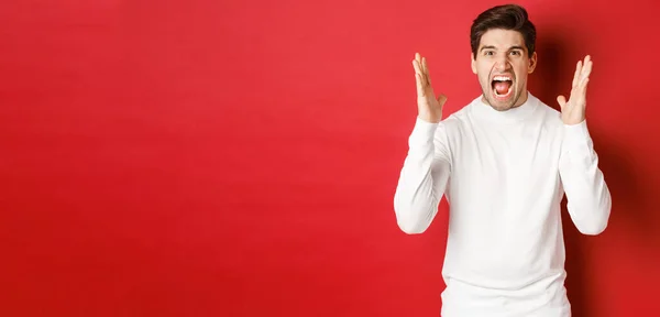 Imagen de hombre frustrado y enojado en suéter blanco, gritando de rabia, enojado con alguien, de pie sobre fondo rojo —  Fotos de Stock