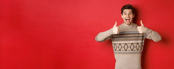 Портрет веселого человека в рождественском свитере, как что-то хорошее, показывая большие пальцы вверх и улыбаясь удивлены, хвалить новогоднюю вечеринку, стоя на красном фоне — стоковое фото