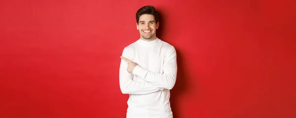Concept of winter holidays, christmas and lifestyle. Portrait of attractive man in white sweater, pointing finger at upper left corner and smiling, showing logo on red background — Stock Photo, Image