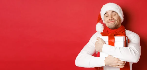 Begreppet vinter semester, jul och livsstil. Bild av härlig kille i Santa hatt och halsduk, krama hans nya år närvarande och leende smickrad, står över röd bakgrund — Stockfoto