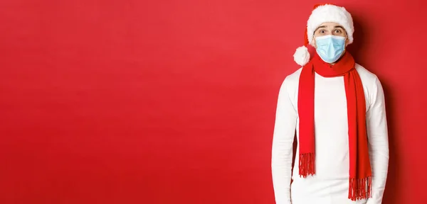 Az újév, a koronavírus és az ünnepek koncepciója. Vidám ember ünnepli a karácsony és a társadalmi távolság, visel orvosi maszk, télapó kalap és sál, álló felett piros háttér — Stock Fotó