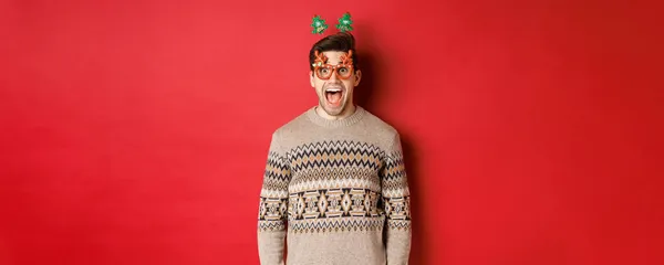 Bild eines überraschten und aufgeregten gutaussehenden Mannes, der eine Partybrille mit Weihnachtspullover, Fallkiefer und ein tolles Neujahrsangebot vor rotem Hintergrund trägt — Stockfoto