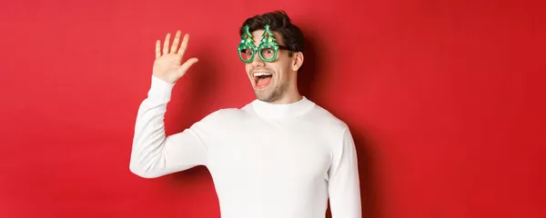 Retrato de homem amigável e alegre em óculos de festa e suéter branco, dizendo olá e olhando para a esquerda, cumprimentando um amigo, em pé sobre fundo vermelho — Fotografia de Stock