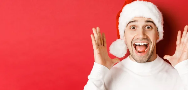 Close-up van gelukkige jonge man in santa hoed, het maken van grote kerst aankondiging, glimlachend verbaasd, staande over rode achtergrond — Stockfoto