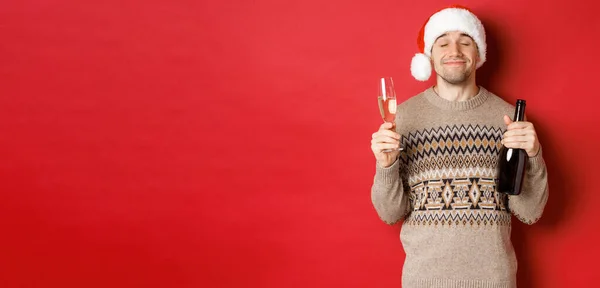Begrip wintervakantie, kerstmis en viering. Afbeelding van tevreden lachende man in kerstmuts en trui, drinken op nieuwjaar, fles champagne en gevuld glas, rode achtergrond — Stockfoto