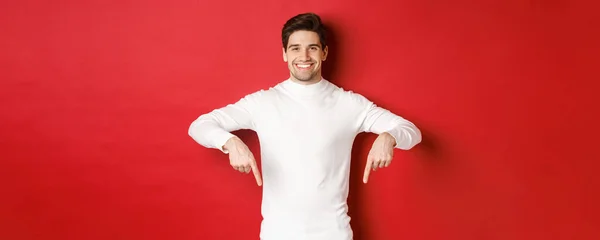 Afbeelding van lachende knappe man in witte trui uitnodigende bezoek pagina, wijzend vingers naar beneden en tonen kerst banner, staande tegen rode achtergrond — Stockfoto