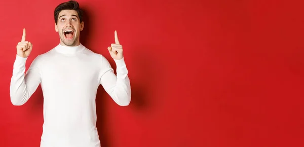 stock image Concept of winter holidays. Image of amazed handsome guy reacting to christmas announcement, pointing and looking up at copy space, standing over red background