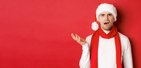 Begreppet jul, vinter semester och fest. Porträtt av förvirrad man i Santa hatt och halsduk, rynkar pannan och ser förbryllad, står över röd bakgrund — Stockfoto