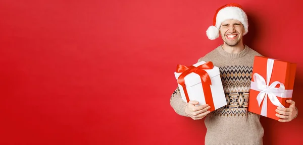 Conceito de férias de inverno, ano novo e celebração. Imagem de jovem feliz e animado gosta de presentes, segurando presentes e sorrindo, vestindo chapéu de Papai Noel e camisola de Natal — Fotografia de Stock