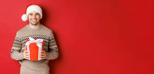 Retrato del hombre guapo sosteniendo un regalo, deseando feliz fiesta, de pie en sombrero de santa y suéter de Navidad sobre fondo rojo —  Fotos de Stock