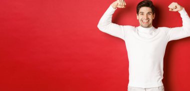 Portrait of smiling handsome man in white sweater, flexing biceps and bragging with strength, show-off strong muscles after workout, standing over red background clipart