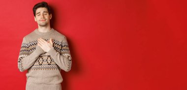 Hayalperest ve mutlu, çekici sakallı bir adamın Noel süveteri giymiş, kalbinin üstünde el ele tutuşmuş, gözleri kapalı gülümseyen, dokunaklı bir şey hatırlayan, yeni yılı kutlayan, kırmızı arka plan