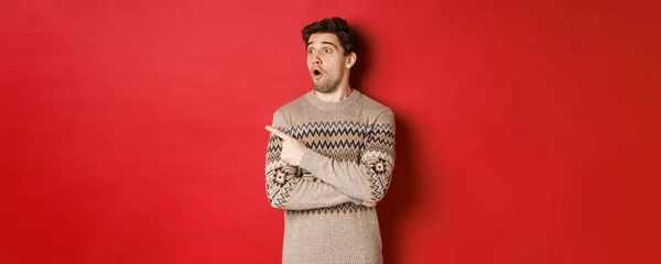 Porträt eines attraktiven kaukasischen Mannes, der Neujahr, Winterurlaub feiert, mit erhobenem Zeigefinger nach links schaut, Weihnachtswerbung zeigt, vor rotem Hintergrund steht — Stockfoto
