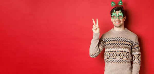 Retrato de hombre divertido y lindo en gafas de fiesta y suéter de Navidad, sonriendo y mostrando señal de paz mientras posando sobre fondo rojo para la publicidad de año nuevo —  Fotos de Stock