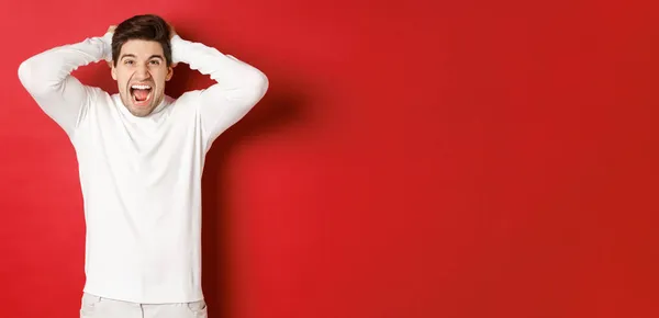 Ritratto di uomo frustrato in maglione bianco, urla di rabbia, si tiene per mano e smorfia, si sente angosciato, in piedi su sfondo rosso — Foto Stock