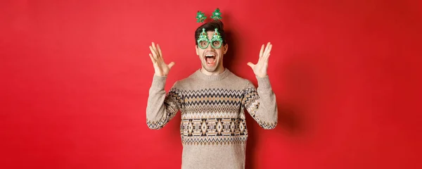 Obraz podekscytowany i zdumiony kaukaski facet w okulary imprezowe, sweter świąteczny, podniesienie rąk do góry i dokonywania dużych ogłoszeń, korzystających z obchodów nowego roku, czerwone tło — Zdjęcie stockowe