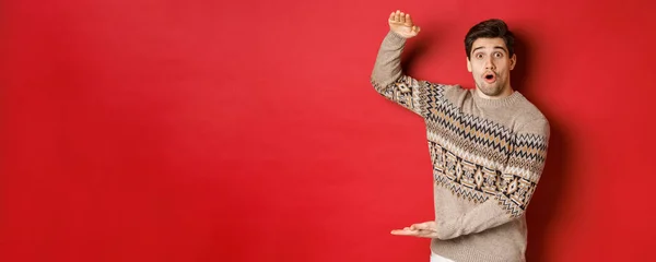 Imagem de homem atraente em camisola de Natal, moldando grande presente de ano novo, mostrando algo grande e incrível, de pé sobre fundo vermelho — Fotografia de Stock