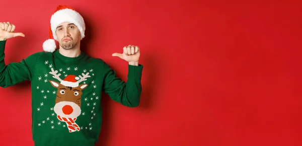 Imagen del joven guapo y confiado en suéter verde y sombrero de santa, apuntándose a sí mismo, celebrando la Navidad, de pie sobre el fondo rojo — Foto de Stock