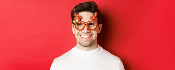 Käsite talvilomat, joulu ja juhla. Lähikuva houkutteleva brunette kaveri puolue lasit, hymyillen ja etsivät onnellinen, seisoo punaista taustaa vasten — kuvapankkivalokuva