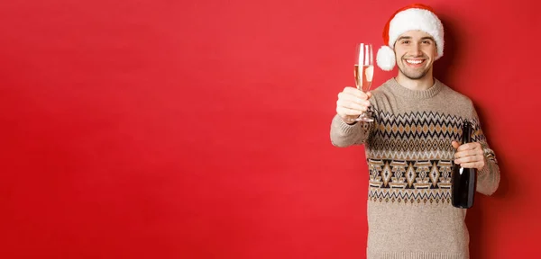 Conceito de férias de inverno, ano novo e celebração. Retrato de homem bonito em santa chapéu e suéter, segurando champanhe, levantando copo e dizendo saúde na festa de Natal — Fotografia de Stock