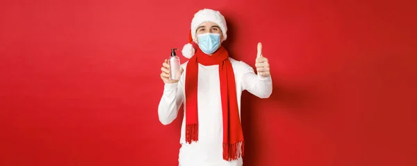 Concept covid-19, karácsony és ünnepek világjárvány idején. Boldog és elégedett férfi télapó kalap és orvosi maszk, mutatja hüvelykujj-up jóváhagyása és ajánló kézfertőtlenítő — Stock Fotó