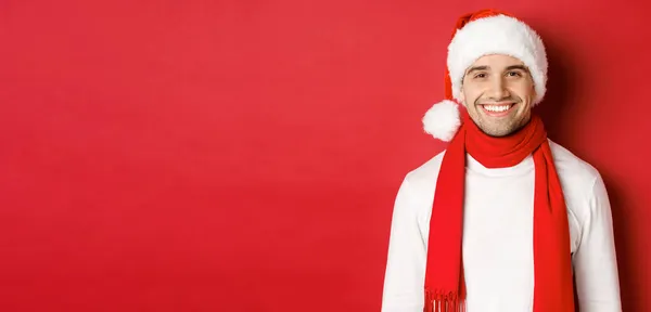 Kış tatili, Noel ve yaşam tarzı kavramı. Kıvırcık saçlı yakışıklı adamın yakın çekimi, eşarplı Noel şapkası takıyor ve gülümsüyor mutlu yıllar diliyor, kırmızı arka plan — Stok fotoğraf