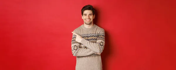 Bild eines gutaussehenden Mannes im Weihnachtspullover, der Neujahr feiert, glücklich lächelt und mit dem Finger auf den Kopierraum in der oberen linken Ecke zeigt, auf rotem Hintergrund stehend — Stockfoto