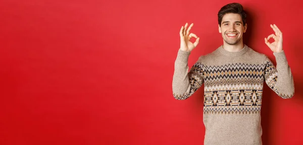 Conceito de celebração de Natal, férias de inverno e estilo de vida. Satisfeito homem bonito em suéter, mostrando sinais ok e sorrindo satisfeito, recomendando algo, fundo vermelho — Fotografia de Stock