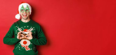 Noel Baba şapkalı ve parti gözlüklü kaygısız yakışıklı adamın portresi, Noel süveteriyle dalga geçiyor, kırmızı arka planda mutlu görünüyor.