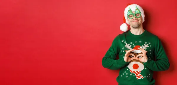 Εικόνα του χαρούμενου χαμογελαστού άνδρα με τα γυαλιά του πάρτι και το καπέλο santa, χαζολογώντας με αστεία χριστουγεννιάτικα πουλόβερ, γιορτάζοντας τις χειμερινές διακοπές, στέκεται πάνω από το κόκκινο φόντο — Φωτογραφία Αρχείου