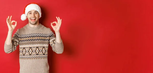 Porträtt av glad och glad stilig man, bär vinter tröja och tomte hatt, visar okej tecken, beröm bra julfest, står över röd bakgrund — Stockfoto