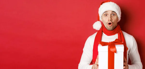 Conceito de férias de inverno, Natal e estilo de vida. Close-up de cara bonito surpreso em santa chapéu e cachecol, olhando espantado e segurando presente de ano novo, de pé sobre fundo vermelho — Fotografia de Stock