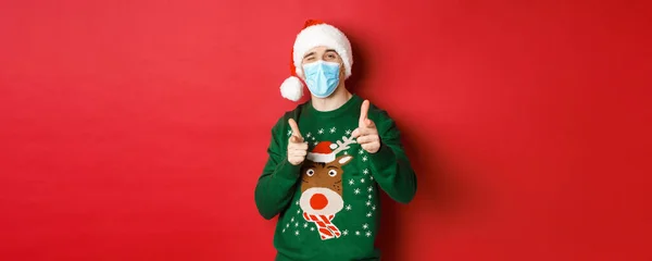 Έννοια του νέου έτους, covid-19 και κοινωνική αποστασιοποίηση. Χαρούμενος άνθρωπος με χριστουγεννιάτικο πουλόβερ, ιατρική μάσκα και καπέλο santa, με τα δάχτυλα στραμμένα στην κάμερα, ευχόμενος καλές διακοπές, στέκεται πάνω από το κόκκινο φόντο — Φωτογραφία Αρχείου