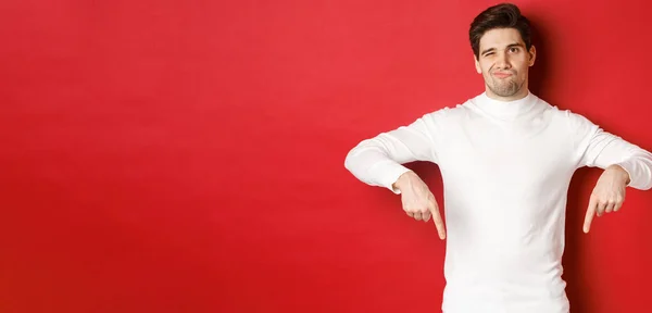 Porträtt av attraktiv, skeptisk ung man i vit tröja, grimma och peka finger ner, oense och ogillar något, står över röd bakgrund — Stockfoto