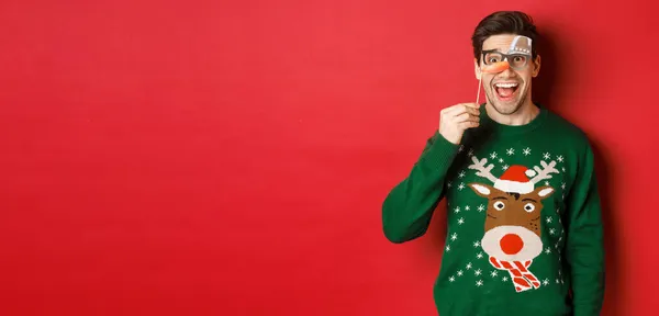 Retrato de un hombre guapo divertido en jersey de Navidad, con una divertida máscara de fiesta y una sonrisa, celebrando las vacaciones de invierno, de pie sobre el fondo rojo — Foto de Stock