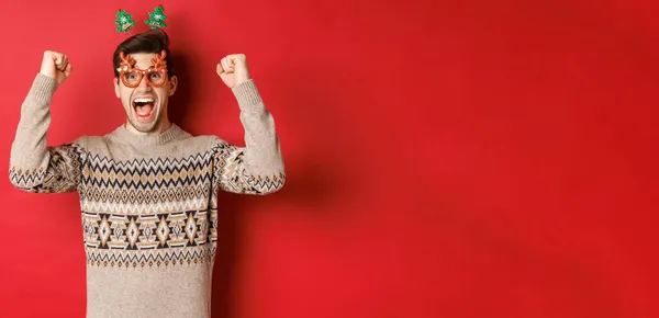 Retrato de hombre emocionado y feliz en gafas de fiesta, suéter de invierno, gritando de alegría y levantando las manos, ganar el premio en Navidad, lograr el objetivo de año nuevo, de pie fondo rojo — Foto de Stock