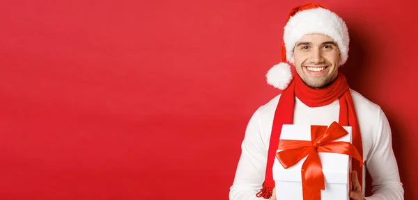 Konzept für Winterurlaub, Weihnachten und Lifestyle. Nahaufnahme eines attraktiven bärtigen Mannes mit Weihnachtsmütze und Schal, der ein Geschenk in der Hand hält, ein Neujahrsgeschenk gibt und lächelt, roter Hintergrund — Stockfoto