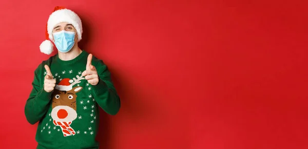 Az új év koncepciója, a kovid-19 és a társadalmi távolság. Vidám férfi karácsonyi pulóverben, orvosi maszk és mikulás kalap, ujjal mutogatva a kamerára, boldog ünnepeket kívánva, vörös háttér felett állva — Stock Fotó