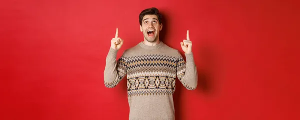 Kuva innoissaan komea kaveri, joka tykkää talvilomien näyttää mainos, etsivät ja sormet ylös hämmästynyt kasvot, seisoo joulun villapaita punaisella taustalla — kuvapankkivalokuva