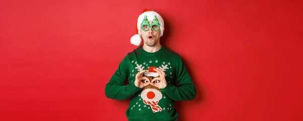 Πορτρέτο του ξέγνοιαστου όμορφου άντρα με το καπέλο και τα γυαλιά του, κοροϊδεύοντας το χριστουγεννιάτικο πουλόβερ του, δείχνοντας χαρούμενος πάνω από το κόκκινο φόντο — Φωτογραφία Αρχείου