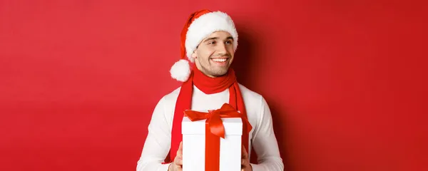Концепция зимних праздников, Рождества и образа жизни. Крупный план привлекательного мужчины в шляпе Санты и шарфе, держащего новогодний подарок, смотрящего направо и улыбающегося, стоящего на красном фоне — стоковое фото