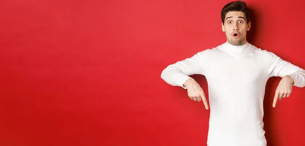 Image d'un gars attrayant surpris en pull blanc, montrant l'annonce, pointant les doigts vers le bas et regardant étonné, debout sur fond rouge — Photo