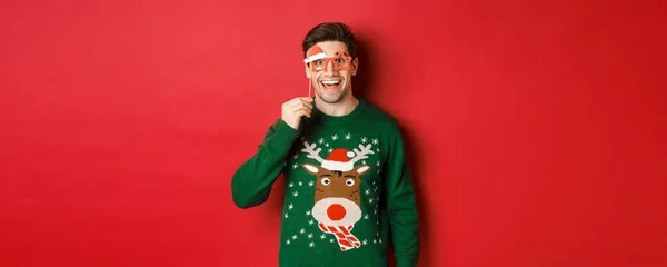 Retrato de hombre guapo sonriente en jersey de navidad y gafas de fiesta, mirando sorprendido y feliz, celebrando año nuevo y divirtiéndose, de pie sobre fondo rojo — Foto de Stock