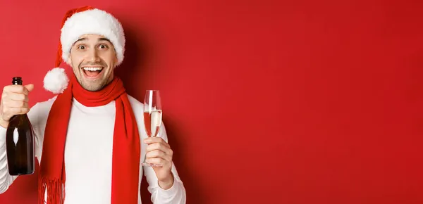 Begrip wintervakantie, kerst en levensstijl. Close-up van vrolijke knappe man met champagnefles en glas, nieuwjaar vieren, over rode achtergrond staan Stockfoto