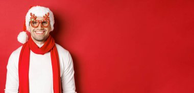 Noel, kış tatili ve kutlama kavramı. Yeni yıl partisinde gözlüklü, gülümseyen, kırmızı arka planda duran neşeli yakışıklı adam.