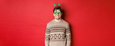 Kış süveteri ve parti gözlükleri içinde mutlu ve yakışıklı bir adamın portresi, yeni yılı ya da Noel 'i kutlamak, kırmızı arka planda dururken gülümsemek.